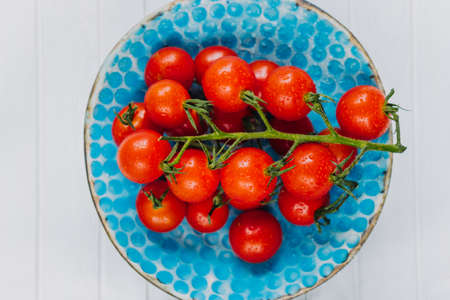 Appetizing ripe cherry tomatoes on a twig in a blue clay plate on a white wooden background. Fresh and healthy vegetarian food.の写真素材
