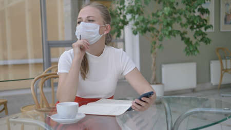 blonde businesswoman with paper notebook and mobile phone takes off medical mask to drink coffee at glass table in cozy cafeの写真素材
