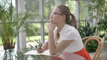 elegant blonde businesswoman in white t-shirt writes in paper notebook with pen sitting at table in cafe with tropical pot plantsの写真素材