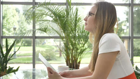 long haired blonde businesswoman with glasses surfs internet with contemporary tablet sitting at table in cafe with pot plantsの写真素材