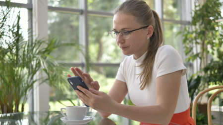positive woman in glasses with ponytail surfs internet with mobile phone drinking coffee at table in...