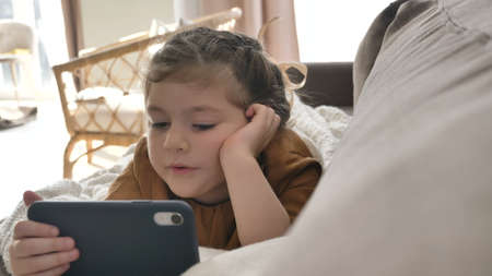 Adorable little girl watches cartoons on modern smartphone lying on soft comfortable sofa in light living room close viewの写真素材