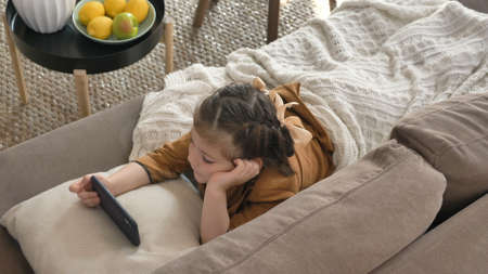 Cute little girl with bows covered with white plaid watches cartoons on cellphone lying on sofa in light living room closeupの写真素材