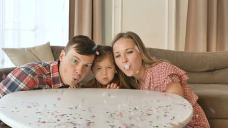 Joyful family of mother father and little daughter blow off confetti from white table in contemporary living room slow motionの写真素材