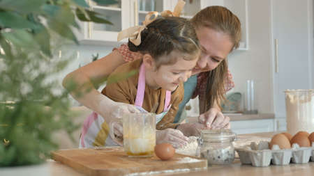 Cheerful young mommy and daughter have fun with fresh white flour cooking pie at wooden table in contemporary light kitchenの写真素材