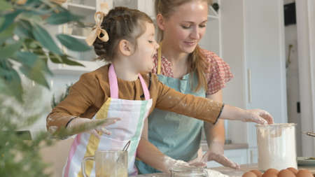 Cheerful young mommy and daughter have fun with fresh white flour cooking pie at wooden table in contemporary light kitchenの写真素材