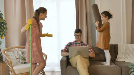 Cute little girl beats daddy with pillow and woman with mop talks to husband working on laptop on sofa at quarantine lock-downの写真素材