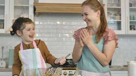 Positive little girl and young mother in aprons play with white flour cooking delicious cake together in contemporary kitchenの写真素材