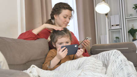 Positive young mother strokes head of little girl holding mobile phones sitting under knitted plaid on sofa in room closeupの写真素材