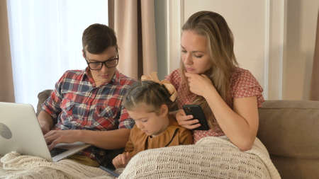 Young family of mother father and little daughter use different contemporary gadgets on sofa in elegant light living roomの写真素材