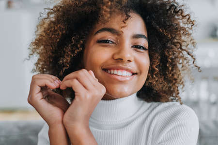 Cheerful model with perfect dark skin and white teeth hols hands in heart shape near face smiling widely at home close viewの写真素材