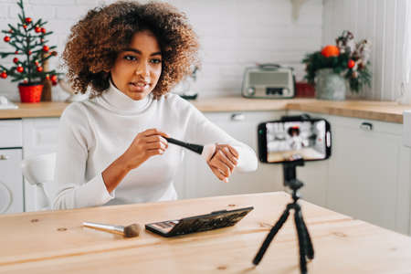Dark skinned blogger holding professional makeup brush and showing tone on hand sitting against...