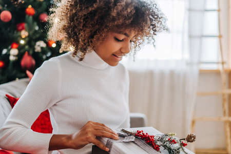 Young woman with dark skin and kinky hair opening present box and smiling widely against decorated Christmas tree closeupの写真素材