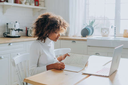 Student girl with trendy kinky hair sitting against notebook and writing notes in copybook enjoying online education closeupの写真素材