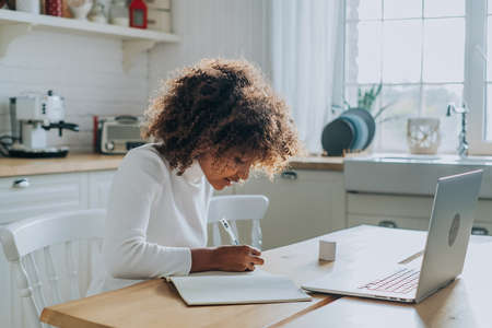 Student girl with trendy kinky hair sitting against notebook and writing notes in copybook enjoying online education closeupの写真素材