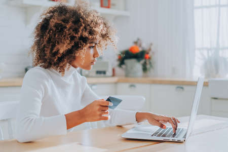 Lady with perfect dark skin holding credit card and inserting bank information for online shopping sits near laptop closeupの写真素材