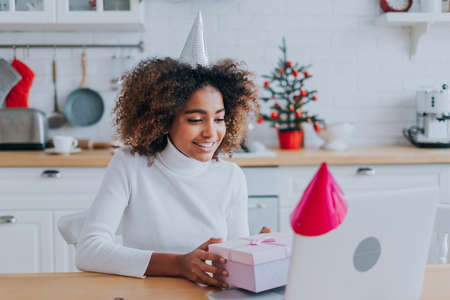 Smiling African lady with kinky hair holds decorated present box near notebook monitor sitting at modern kitchen closeupの写真素材