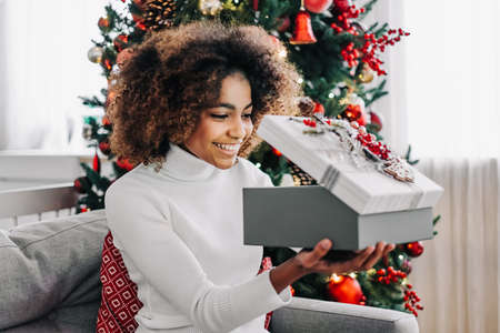 Young woman with dark skin and kinky hair opening present box and smiling widely against decorated Christmas tree closeupの写真素材