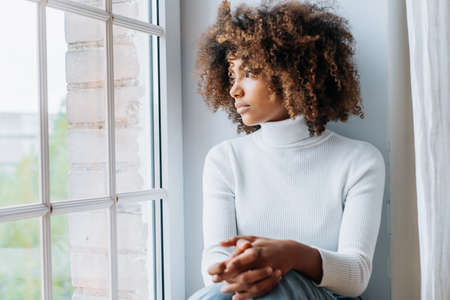 Thoughtful young woman with dark skin and modern kinky hair sits on white windowsill looking outside window close viewの写真素材