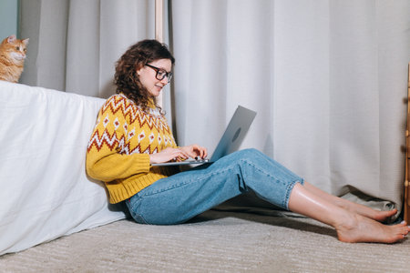Woman in a yellow sweater and glasses works at home at a laptop sitting on carpet with her red cat, remote work from home, freelance.の写真素材