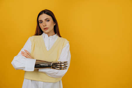Portrait of a beautiful young woman with a silver bionic prosthetic arm in a white shirt on a yellow...