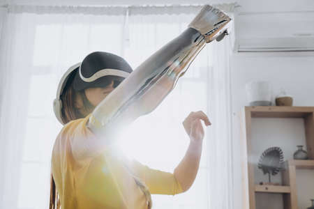 A young woman with a bionic hand in virtual reality glasses has her hands up and touches the air of...