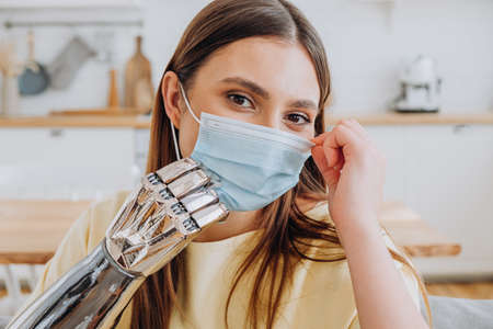 Positive blonde lady puts on blue disposable protective mask by modern metal bionic prosthesis arm in light kitchen closeupの写真素材