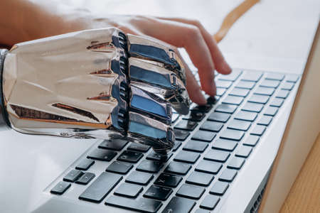 Young woman uses modern metal bionic arm to type on laptop keyboard sitting at wooden table in light room extreme close viewの写真素材