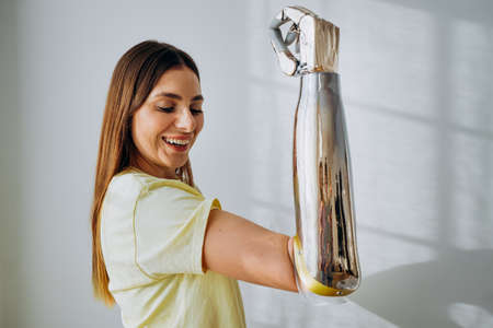 Positive woman in t-shirt does winner gesture with contemporary bionic prosthesis arm standing at white wall in room closeupの写真素材