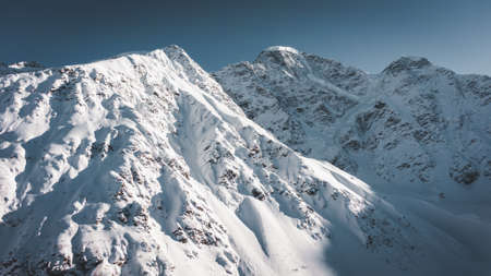 Amazing old mountains with steep slopes covered with shiny snow and shadows under blue sky in sunny winter morning aerial viewの写真素材