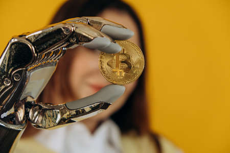 Lady with modern shiny metal bionic arm holds large golden bitcoin posing for camera on yellow background extreme close viewの写真素材