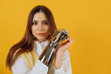 Long haired blonde young woman in white shirt and vest shows contemporary bionic arm standing on bright yellow backgroundの写真素材