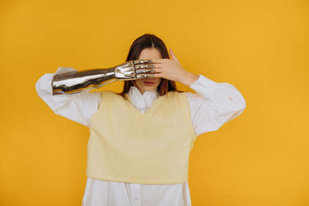 Pretty young lady in shirt and vest with contemporary bionic prosthesis arm closes eyes by palms on yellow background closeupの写真素材