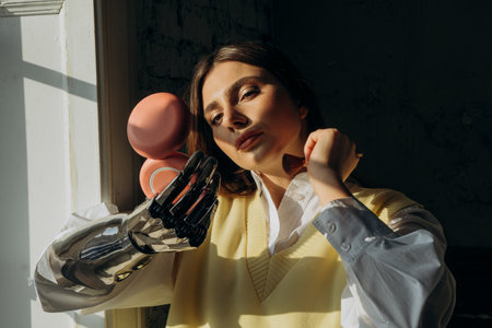 Attractive woman with a bionic prosthetic arm applies makeup with a brush standing at the window and looks in a small mirrorの写真素材