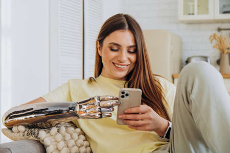A happy woman with a bionic prosthetic arm sits on the couch with a phone and communicates online with friends or surfs the Internet or makes online purchasesの写真素材