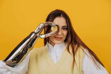 Lady with shiny bionic arm holds golden bitcoin on yellowの写真素材