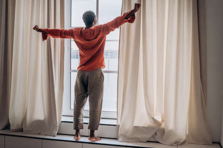 African man stretches after waking up standing at the window and looks into the distanceの写真素材