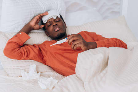 Sick African man takes his temperature while lying in bed with a cooling compress on his headの写真素材