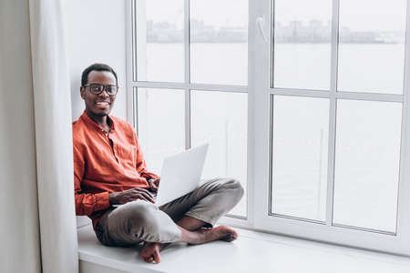 A young man African businessman with glasses is sitting at a laptop on the windowsill of a house looking at the camera and smilingの写真素材