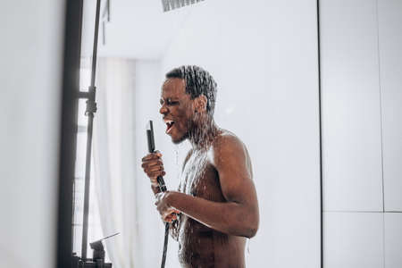 Happy African-American man sings in the shower into an imaginary microphone under a stream of waterの写真素材
