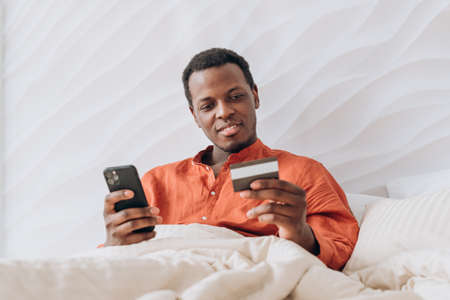 African-American guy holds smartphone and credit card in bedの写真素材