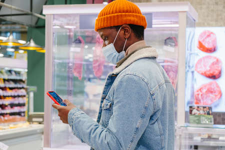 African-American man in mask uses phone in supermarketの写真素材
