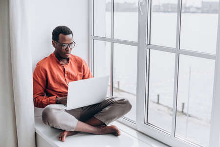 Black man surfs internet on laptop sitting on sillの写真素材