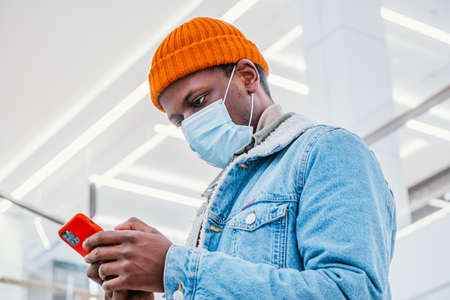 African-American guy in mask uses mobile phone in hallwayの写真素材