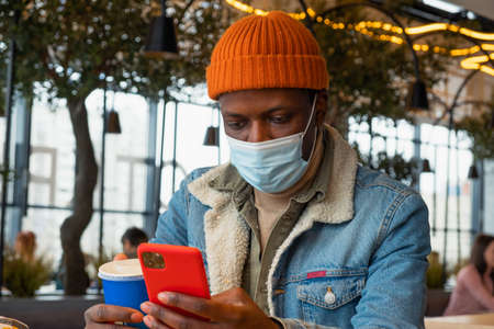 Black man in face mask surfs internet with phone in cafeの写真素材