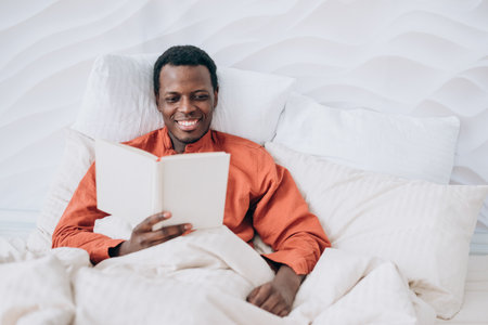 Black man in pajamas reads book lying in comfortable bedの写真素材