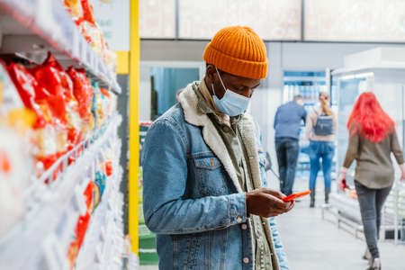 Black man with mask uses smartphone by supermarket racksの写真素材