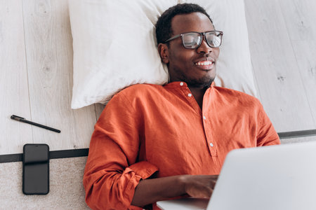 Cheerful black male student works on laptop lying on floorの写真素材