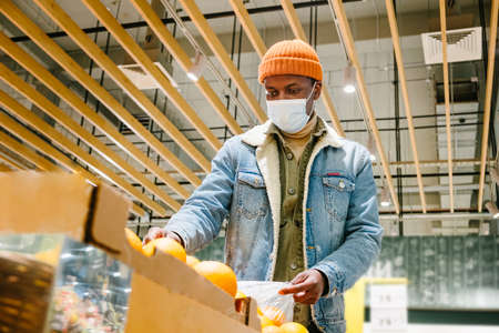 African-American person in mask takes oranges in supermarketの写真素材