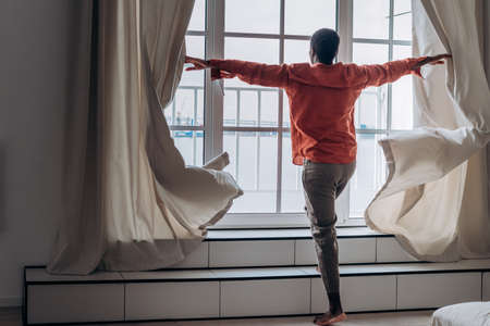 African-American man in orange shirt opens curtains in roomの写真素材